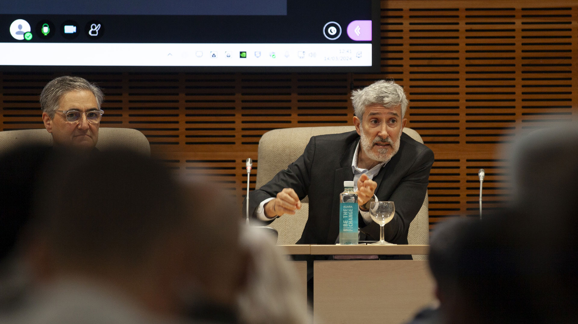 Ein Mann sitzt an einem Tisch mit einem Bildschirm im Hintergrund.